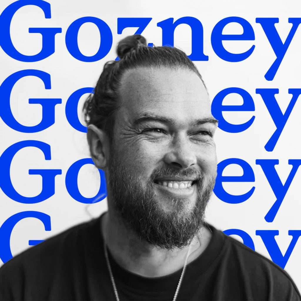 Bearded man with a topknot smiling, shown in black and white, in front of a white background with the word “Gozney” repeatedly printed in large blue letters.