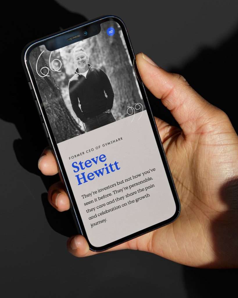 A hand holds a mobile phone displaying a profile of Steve Hewitt, former CEO of Gymshark. The screen shows a black-and-white photo of him and a quotation about investors' qualities, with his name in large blue text.