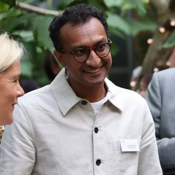 A man with short curly hair and glasses, wearing a light-coloured jacket and name tag, smiles whilst talking to a woman at an outdoor event with greenery in the background.