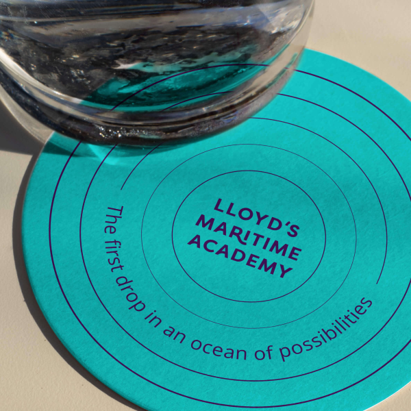 A round turquoise coaster with concentric circles, featuring the text LLOYDS MARITIME ACADEMY and The first drop in an ocean of possibilities. A glass of water is partly placed on the coaster.