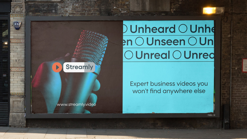A billboard shows a close-up of a hand holding a microphone on the left and blue text on the right reading Unheard, Unseen, Unreal. Below, it says, Expert business videos you won’t find anywhere else.