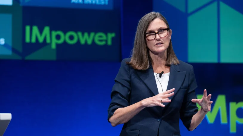 A woman wearing glasses and a navy blazer speaks on stage with “IMpower” displayed on a large blue and green screen behind her. She gestures with her hands whilst presenting.