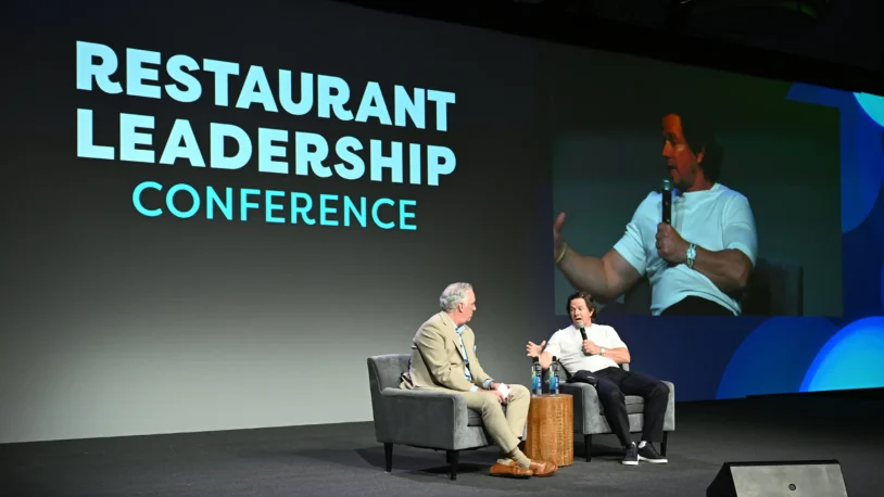 Two men sit on stage in armchairs, having a discussion at the Restaurant Leadership Conference. One holds a microphone whilst the other listens. Their conversation is projected on a large screen behind them.