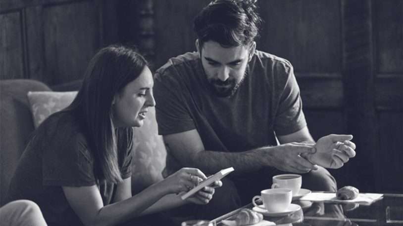 A woman and a man from a B2B brand sit closely on a sofa, looking at a mobile in the woman's hand. Coffee cups and pastries rest on the glass table as they engage in a serious conversation, perhaps about their digital agency's next project.
