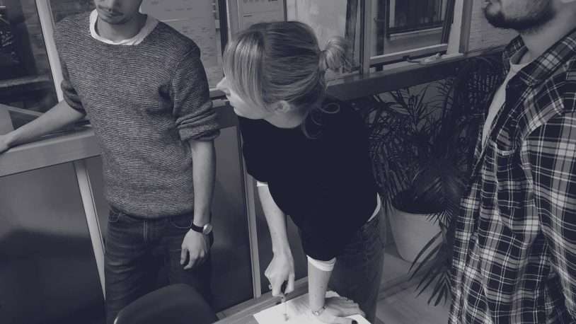 Three people stand around a table in a digital agency office. One woman points at papers as the group discusses strategy, collaborating on ideas for a leading B2B brand.