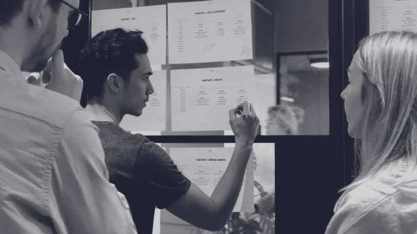Three people stand in front of a glass wall with papers attached, as one person writes or points to a document whilst the others observe and discuss strategies for their B2B brand and digital agency.