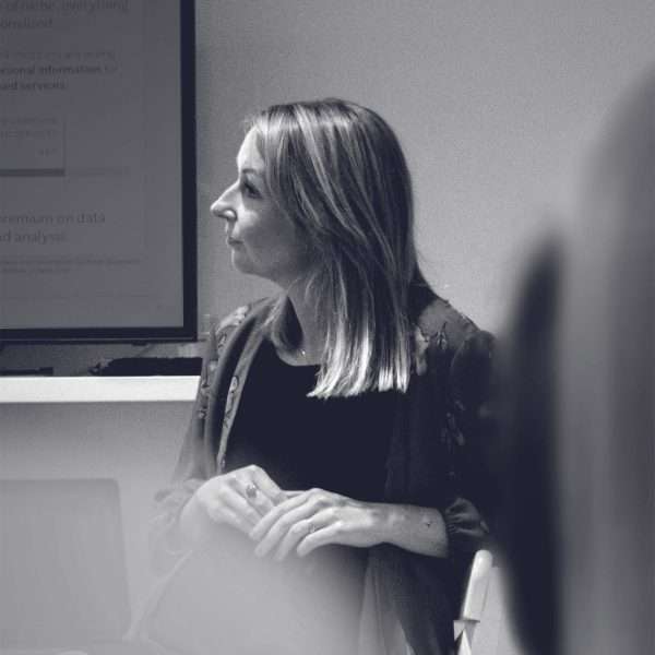 A woman with straight, shoulder-length hair sits and looks to the left in a softly lit room. She wears a patterned jacket over a dark shirt. A blurred figure and a presentation screen are visible in the background.