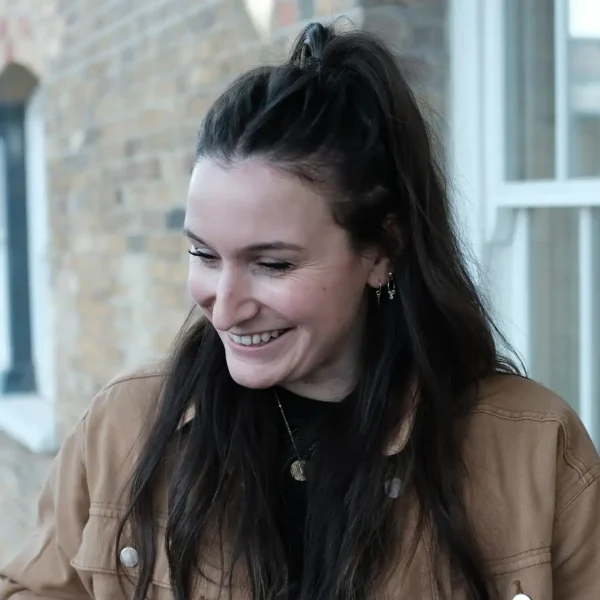 A woman with long dark hair, wearing a brown jacket and a black top, smiles whilst looking down. She stands outside in front of a brick wall and a white-framed window.