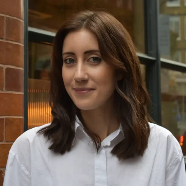 A woman with shoulder-length brown hair and a white button-up shirt stands in front of a brick wall and window, smiling softly at the camera.