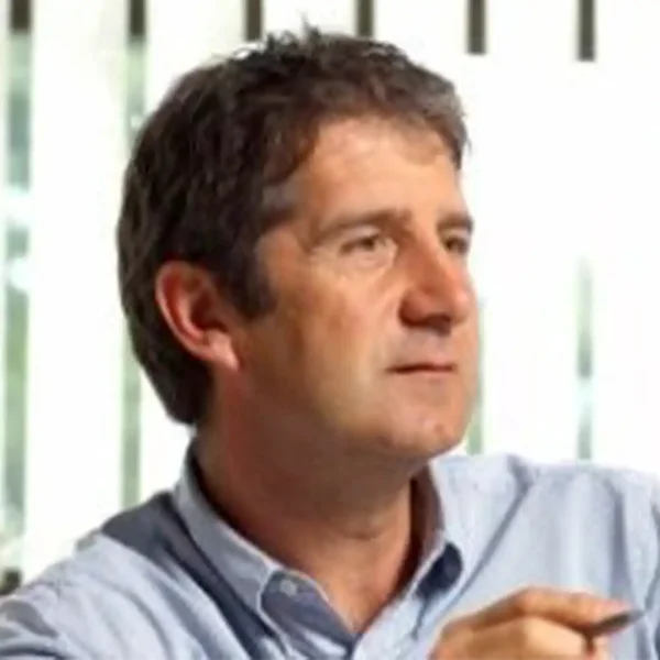 A middle-aged man with short, dark hair wearing a light blue collared shirt sits indoors at a B2B brand and digital agency, looking to the side. Vertical blinds and natural light are visible in the background.