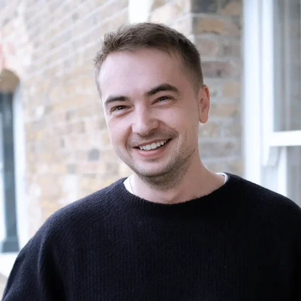 A young man with short brown hair and a trimmed beard smiles while standing outside in front of a brick wall and window, wearing a black jumper.