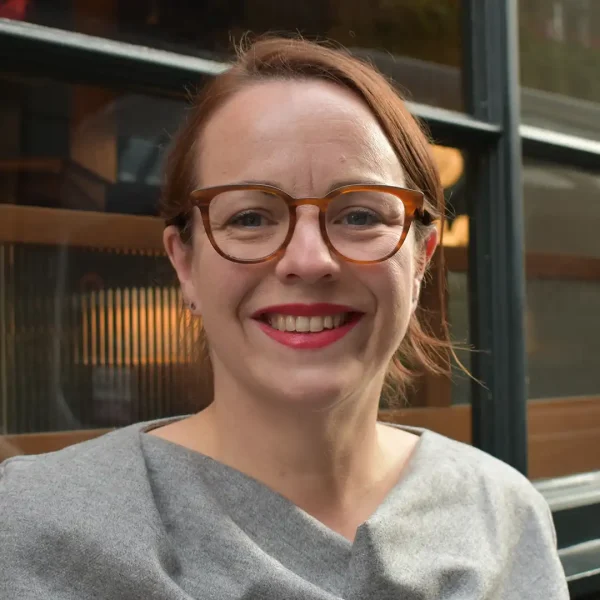 A smiling woman with brown hair, wearing glasses and a light grey top, sits in front of a window with a dark frame and wooden panels.