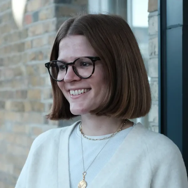A young woman with straight brown hair and black glasses is smiling. She is wearing a cream-coloured jumper, layered necklaces, and standing in front of a brick wall and a window.