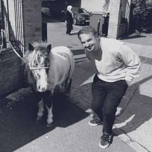 A man in a light jumper and dark trousers stands smiling next to a small, spotted pony on a paved path. A person is visible by parked cars in the background. The image is in black and white.