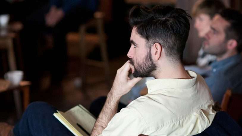 A man with dark hair and a beard sits in profile, holding a notebook and pen, looking thoughtful. Other people are seated nearby, slightly out of focus in the background. The setting appears casual and indoors.