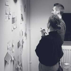 Two people stand facing a wall covered with sticky notes, discussing and reviewing ideas. One person holds a pen and notebook, while both appear engaged in a brainstorming or planning session.