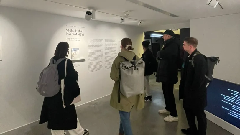 Five people with rucksacks stand in a gallery, reading wall text about the artist Sasha Huber’s exhibition titled YOU NAME IT, displayed on a white wall under spotlights.