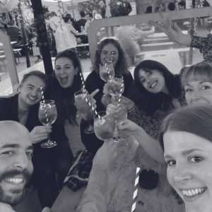 A group of seven people sit around a table outdoors, smiling and holding up drinks in a celebratory toast. The atmosphere is lively, with other people visible in the background. The image is in black and white.