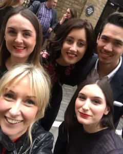 Five people, three women in front and one woman and one man behind, smile at the camera outdoors on a street with brick buildings in the background.