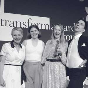 Four people pose and smile together at an awards event. One person holds a trophy. The background displays the word transform. Everyone appears happy and celebratory.