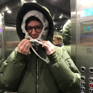 A person in a green puffy jacket with the hood tightly drawn round their face stands in a lift, holding the hood strings. Their reflection is visible in the lift’s mirrored walls.