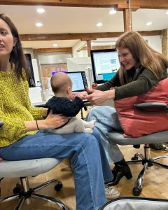 Two women sit in office chairs, one holding a baby on her lap while the other reaches out to interact with the baby. Computer monitors and office equipment are visible in the background.