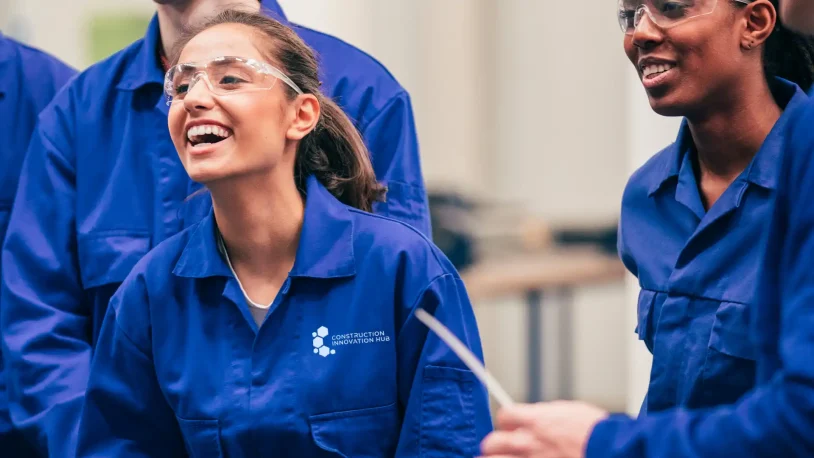 Two people wearing blue overalls and safety glasses smile and laugh whilst working together indoors. The overalls have a Construction Innovation Hub logo on the chest.