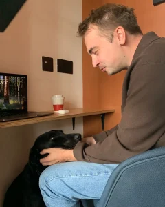 A man sits at a desk, gently holding a black dog’s head and looking at it lovingly. A laptop, coffee cup, and earphones are on the desk, and the dog is sitting under the desk looking up at the man.