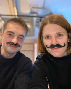 A man and a woman, both wearing black fake moustaches and dark jumpers, sit indoors and smile at the camera. The background is softly blurred, showing some ceiling pipes and bright lighting.
