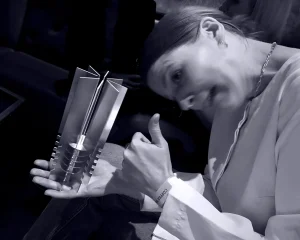 A person smiles broadly and gives a thumbs-up whilst holding a modern, geometric metal trophy. The scene appears celebratory and is lit with dramatic, low lighting.