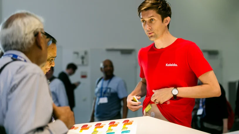 A man in a red Kaleido shirt stands at a table with colourful objects, talking to two older men, whilst people are visible in the background at what appears to be an event or conference.