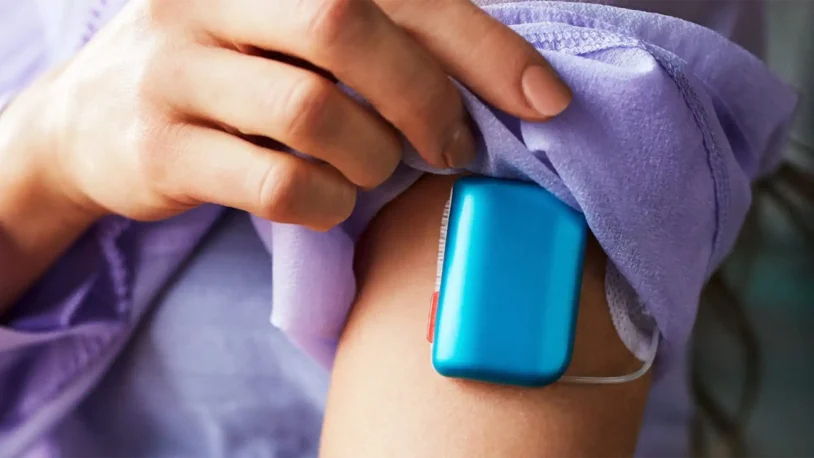 A person lifts their sleeve to reveal a blue medical device attached to their upper arm, likely a wearable injector or insulin pump.