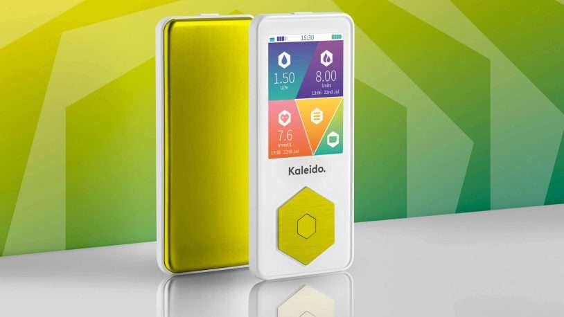 A modern medical device with a touchscreen displaying colourful health stats stands upright next to its yellow cover. The background features geometric green and yellow patterns. The device is labelled Kaleido.