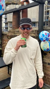 A smiling man in a white long-sleeve shirt and black cap holds a drink can. He wears pixelated deal with it sunglasses. Colourful balloons and a brick wall are in the background at an outdoor gathering.