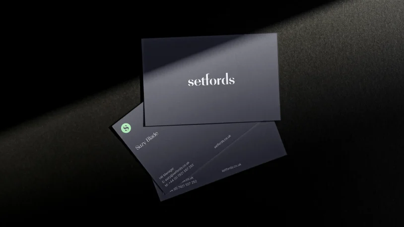 Three dark business cards on a black surface under a beam of light. The top card displays the word setfords in white; the other two cards show contact details and a green logo.