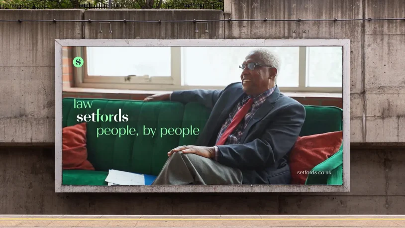 A billboard shows an older man in glasses and a suit sitting on a green sofa, smiling. Text reads: law setfords people, by people. The setfords.co.uk website appears in the corner.