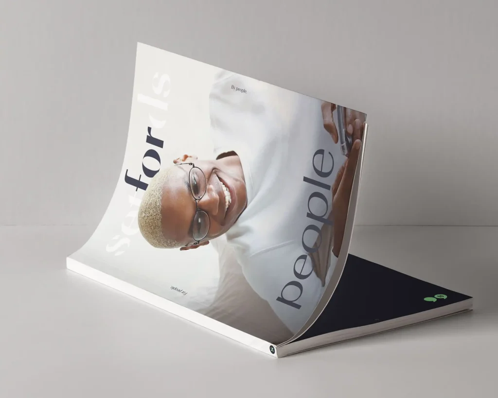 A magazine lies open on a flat surface. Its cover features a smiling person with short blond hair and glasses, wearing a white shirt. The words “Saxfords” and “people” are printed in large letters across the cover.