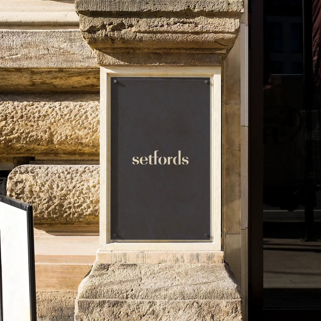 A black sign with the word setfords in white lowercase letters is mounted on a textured stone wall outside a building.