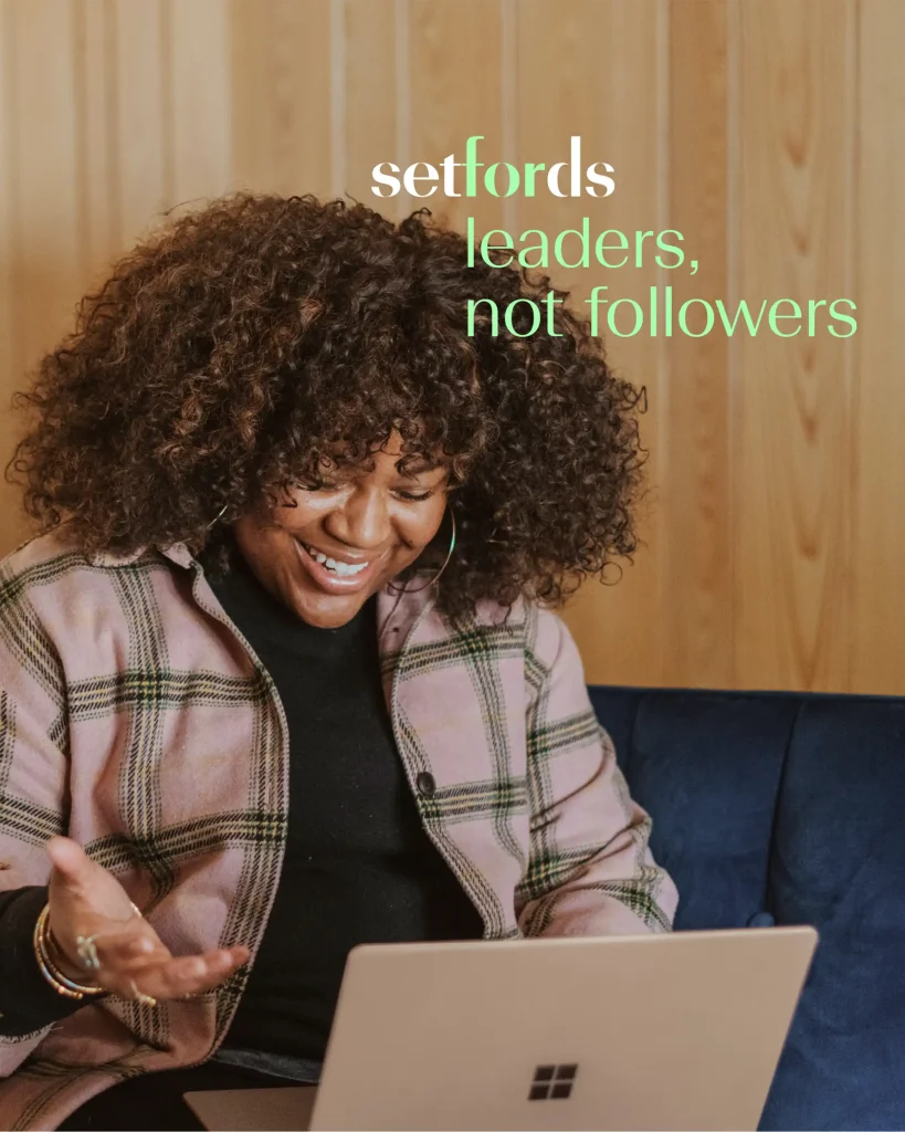 A woman with curly hair smiles and gestures while looking at a laptop. Text on the image reads: setfords leaders, not followers. The background features wood panelling and a blue sofa.
