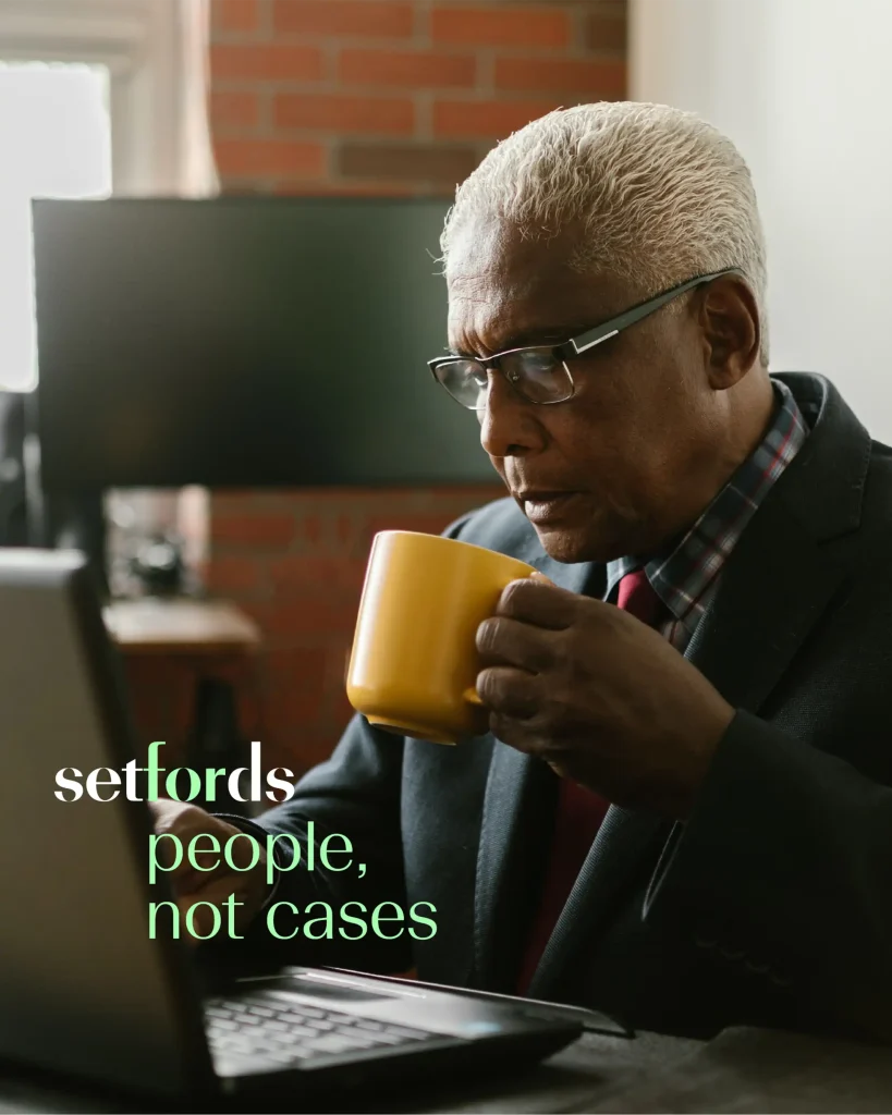 An older man with grey hair and glasses sits at a desk, holding a yellow mug and looking at a laptop. The text reads: “setfords people, not cases.” A computer monitor and brick wall are in the background.