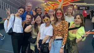 A group of people pose together outdoors at an event, smiling and making peace signs. Colourful lights create a vibrant background. Most are wearing casual clothes, and some have sunglasses or festival accessories.