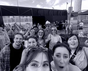 A group of people smiling and posing for a selfie at an outdoor event, with others in the background. The sky is partly cloudy and there are buildings and fences visible behind them.