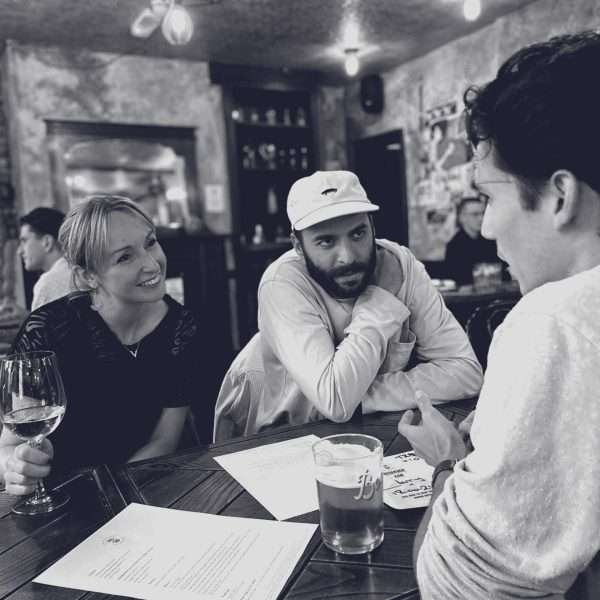 Three people sit at a wooden table in a cosy bar, engaged in conversation. One woman holds a glass of wine, while the two men listen intently. Papers and drinks are on the table. The scene is relaxed and social.