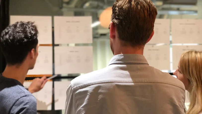 Three people from a B2B brand and digital agency stand facing a wall covered with sheets of paper, discussing or analysing the documents. The photo is taken from behind them, focusing on their backs and the papers.