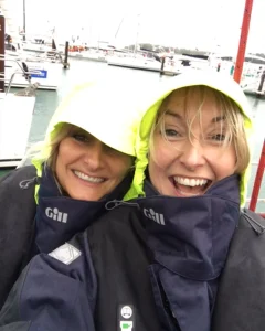 Two women in navy waterproof jackets with yellow hoods smile at the camera whilst standing on a quay with boats and water in the background on a cloudy day.