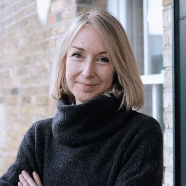 A woman with shoulder-length blonde hair and a black polo-neck jumper stands in front of a brick wall, smiling softly with her arms folded.