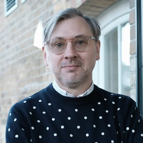 A man with fair hair and glasses, wearing a navy blue jumper with white polka dots, stands in front of a brick building and looks at the camera, smiling slightly.