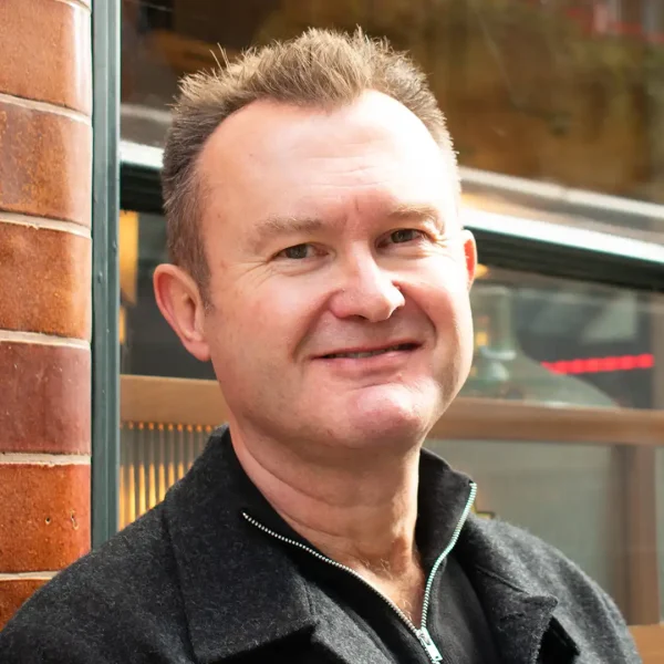 A middle-aged man with short brown hair smiles whilst standing outside near a brick wall and a window, wearing a dark zip-up jacket.