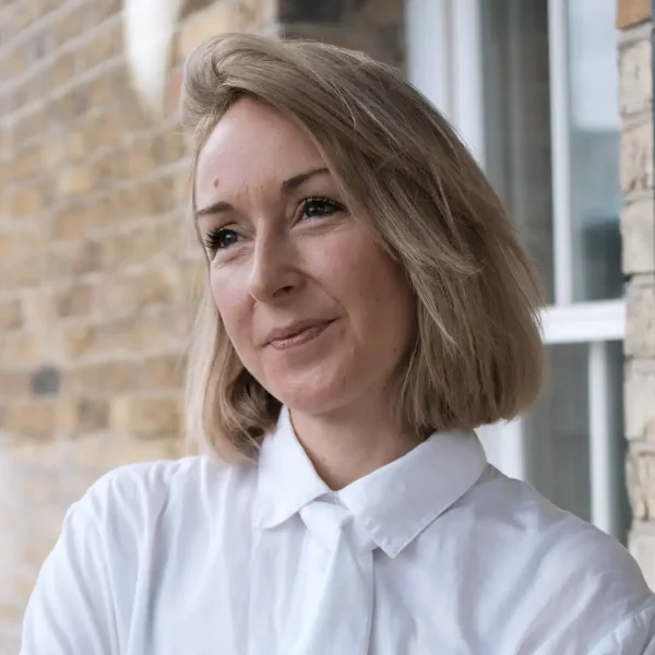 A woman with short blonde hair wearing a white collared shirt stands in front of a brick wall, smiling softly and looking slightly to the side.