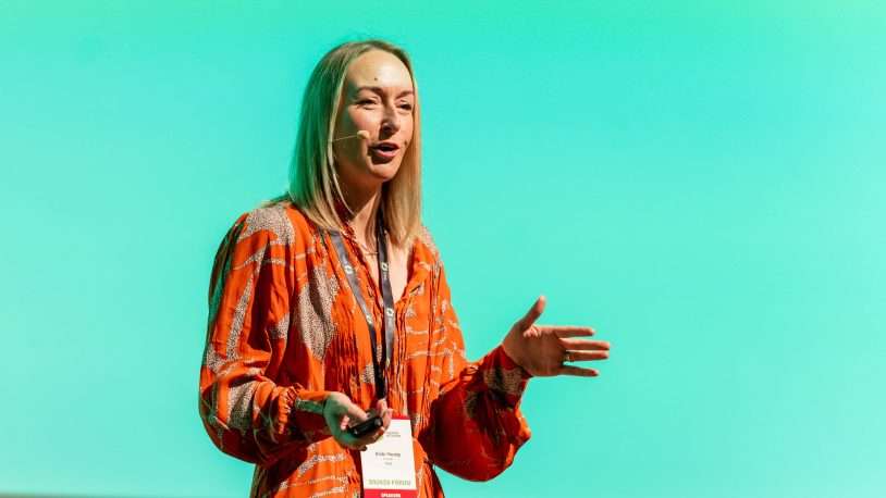 A woman wearing a red patterned dress and a headset microphone is speaking on stage, gesturing with her hands, against a bright turquoise background. She has a conference badge round her neck.